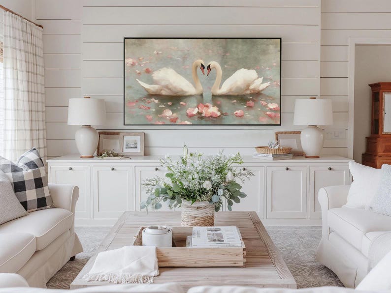 May include: A living room scene featuring a large framed artwork of two swans forming a heart shape with their necks. The artwork hangs above a white cabinet with two matching lamps. A coffee table with a floral arrangement and two white sofas complete the room.