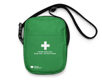 May include: A green crossbody bag with a black adjustable strap and buckle. The front features a white cross symbol, the text "Handle with Care Keep Cool - Do Not Freeze," and a heart icon with the text "ORGAN TRANSPORT" printed in white.