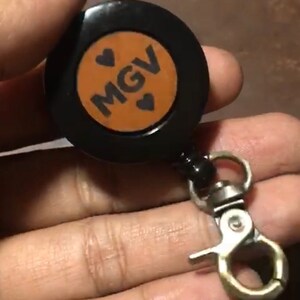 May include: A black retractable badge holder with a brown leather insert. The insert has the text "MGV" with two hearts on either side of the text. The badge holder is attached to a silver swivel clip.