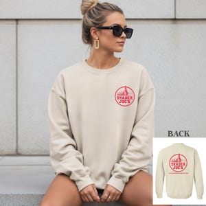May include: A cream-colored sweatshirt with a red Trader Joe's logo on the front. The logo is a circular design with the words "Trader Joe's" and an image of a tower. The back of the sweatshirt is visible in a smaller image.