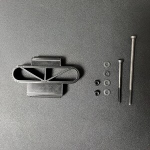 May include: A set of black 3D-printed components, including a rectangular base with an attached oval structure, washers, nuts, and screws. The parts are arranged on a dark grey surface.