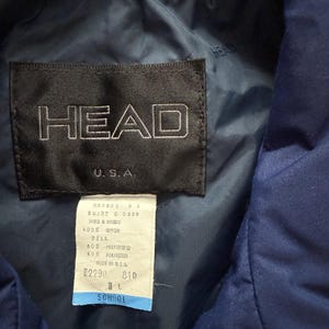 May include: Close-up of a navy blue jacket label with the brand name "HEAD" in white lettering. The label also includes "U.S.A." and a smaller tag with product information. The jacket appears to be made of nylon and polyester.