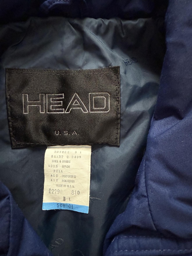 May include: Close-up of a navy blue jacket label with the brand name "HEAD" in white lettering. The label also includes "U.S.A." and a smaller tag with product information. The jacket appears to be made of nylon and polyester.