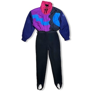 May include: A vintage ski suit featuring a color-blocked jacket in purple, blue, black, and pink, paired with black stirrup pants. The jacket has a high collar and long sleeves. The suit is displayed on a white background.