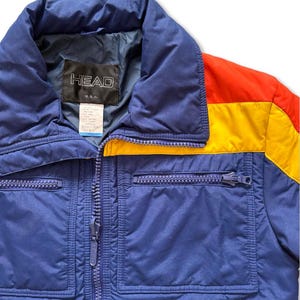 May include: A vintage blue ski jacket with a full zipper closure and two front pockets. The jacket features a color block design with red and yellow accents on the right shoulder. The brand "HEAD U.S.A." is visible on the label.