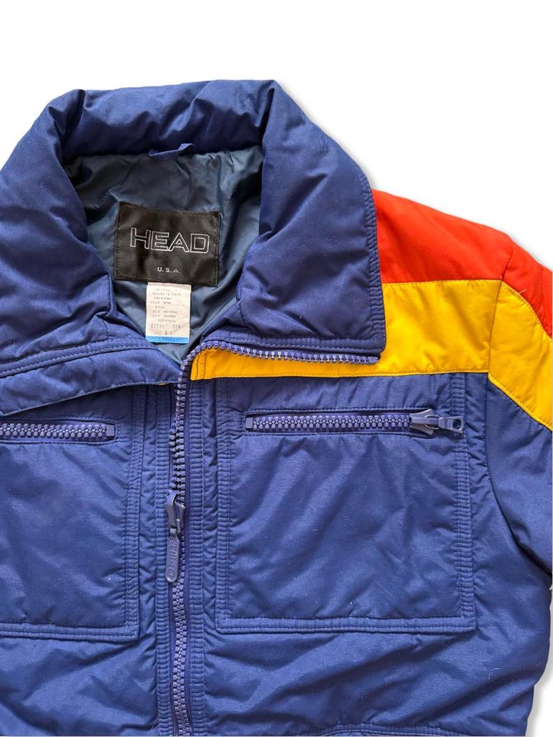 May include: A vintage blue ski jacket with a full zipper closure and two front pockets. The jacket features a color block design with red and yellow accents on the right shoulder. The brand "HEAD U.S.A." is visible on the label.