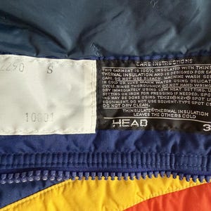 May include: Close-up of a section of a colorful winter jacket. The jacket features a blue, yellow, and orange color scheme with a zipper. There are two white tags with numbers and the letter "S". A black tag with care instructions and the "HEAD" and "3M" logos is also visible.