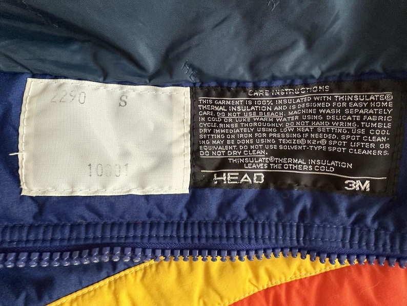 May include: Close-up of a section of a colorful winter jacket. The jacket features a blue, yellow, and orange color scheme with a zipper. There are two white tags with numbers and the letter "S". A black tag with care instructions and the "HEAD" and "3M" logos is also visible.