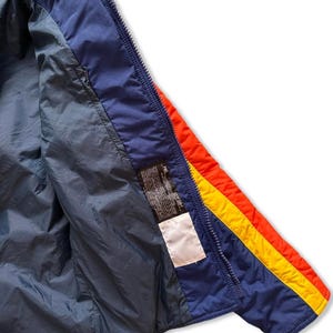 May include: A vintage blue, red, and yellow ski jacket. The jacket has a blue interior and a zipper closure. The exterior features a blue base with red and yellow stripes on the sleeve. A label is visible inside the jacket.