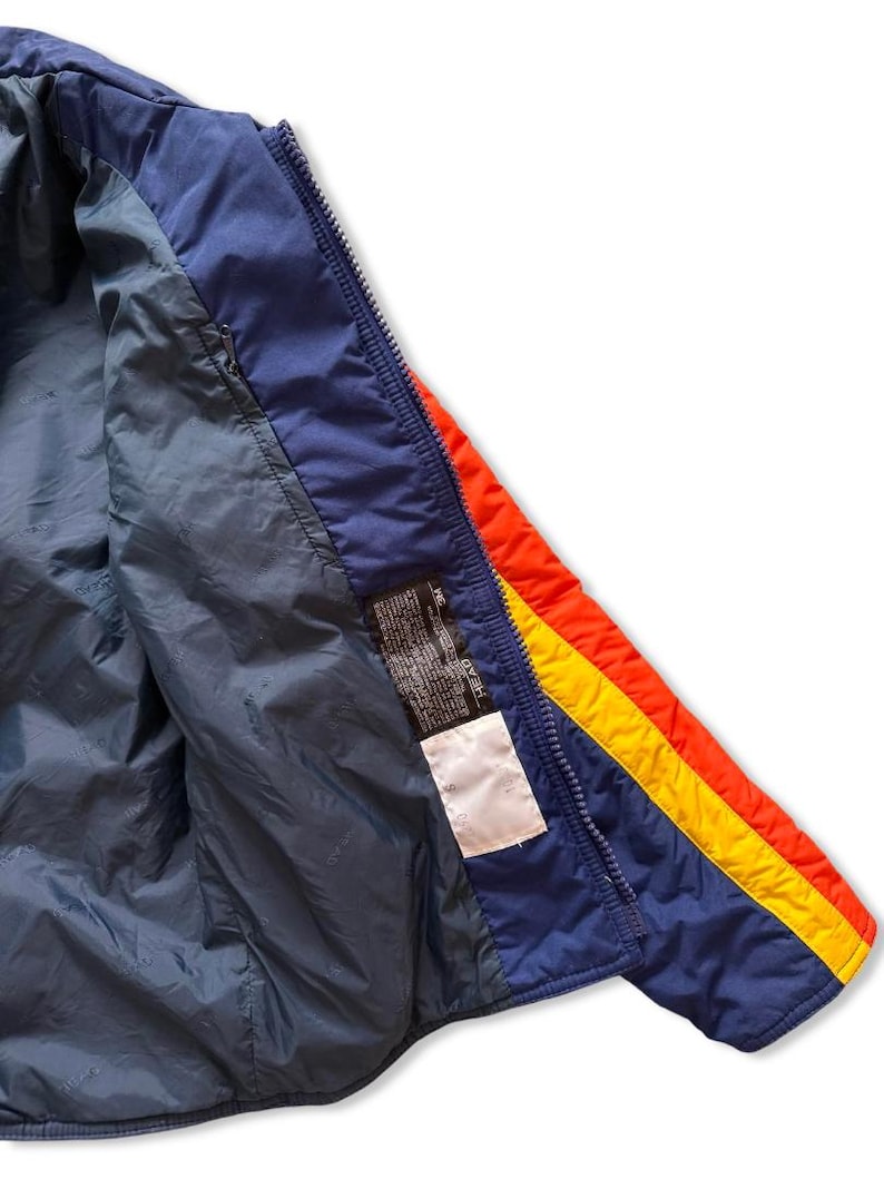 May include: A vintage blue, red, and yellow ski jacket. The jacket has a blue interior and a zipper closure. The exterior features a blue base with red and yellow stripes on the sleeve. A label is visible inside the jacket.