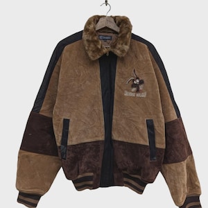 Peut inclure: Une veste en daim marron et marron foncé avec un col en fausse fourrure, des panneaux latéraux noirs, ainsi qu'un ourlet et des poignets côtelés. La poitrine présente un motif brodé d'un personnage avec le texte "UNIVERSE HOLIDAY" en dessous.