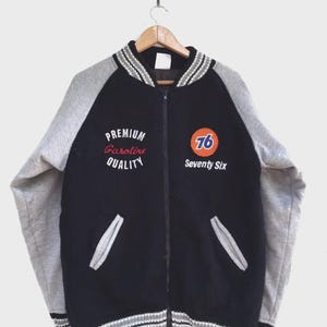 Pode incluir: Casaco universitário vintage preto e cinza com o texto "Premium Gasoline Quality" e "Seventy Six". O casaco tem fecho de correr, mangas cinzentas e detalhes listrados na gola, punhos e bainha.