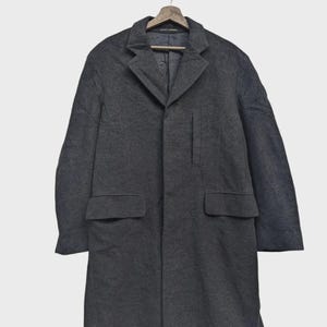 Peut inclure: Un pardessus gris anthracite avec un design classique, avec un revers cranté, deux poches avant et une fermeture à un seul bouton. Le manteau est suspendu à un cintre en bois.