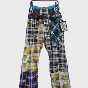 May include: Multi-colored plaid pants with a patchwork design. The pants feature a mix of black, white, blue, green, and yellow plaid patterns. A tag with the text "01.03" hangs from the belt loop. The pants are hanging on a hanger.