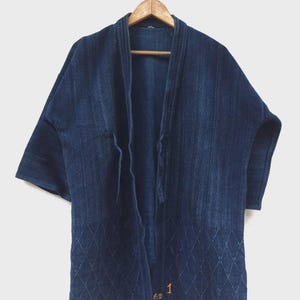 May include: A dark blue, short-sleeved jacket with a diamond pattern on the lower portion. The jacket is hanging on a wooden hanger. The jacket has a textured appearance and a relaxed fit. The number "1" is visible on the lower right side of the jacket.