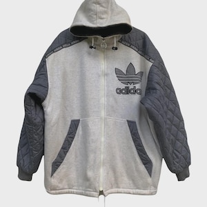 Op de afbeelding: Een grijze en houtskoolkleurige gewatteerde hoodie met ritssluiting en capuchon. Het Adidas-logo staat op de borst. De mouwen en schouders zijn houtskoolkleurig en de body is grijs. Het Adidas-logo is zwart.