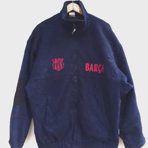 May include: Navy blue fleece jacket with a full zip and a high collar. The jacket displays the word "BARÇA" in red and a red emblem on the chest. A black patch is on the left sleeve.