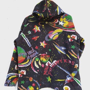 May include: A black hooded sweatshirt with a colorful space-themed pattern. The design includes planets, rockets, stars, and the text "MINX FOR HAPPY KIDS". The sweatshirt has long sleeves and a hood.