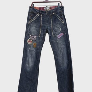 May include: Dark wash denim jeans with decorative patches and distressed details. Features a plaid interior waistband and unique pocket designs. Patches include text that reads "Ocean Breeze" and "Burnze".