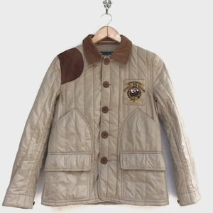 May include: Tan quilted jacket with brown corduroy collar and shoulder patch. Features a button-up front, two front pockets, and a patch with a crest. The jacket is hanging on a wooden hanger.