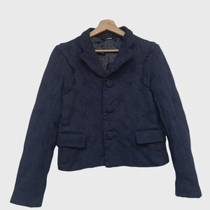 May include: A navy blue blazer with a quilted pattern, featuring a notched collar, three buttons, and two front pockets. The jacket is displayed on a wooden hanger against a white background. The blazer has long sleeves.