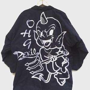 May include: A black jacket featuring a large white graphic of a cartoon devil holding a trident and wearing a cross necklace. The text "OH A DEVIL" is printed in white to the left of the character. The jacket has long sleeves and a collar.