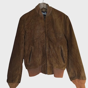 May include: A brown leather jacket with a zip-up front, ribbed cuffs, and a ribbed hem. The jacket has two front pockets and a collar. The brand name "Schott" is visible on the collar. The jacket is hanging on a wooden hanger.