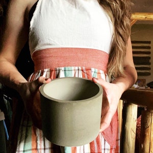 May include: A person is holding a gray concrete planter. The planter is cylindrical and has a smooth surface. The person is wearing a red and white plaid dress.