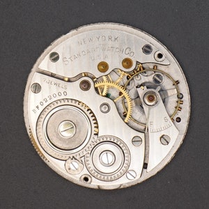 May include: Close-up view of a silver pocket watch movement with gold gears. The watch face reads "NEW YORK STANDARD WATCH CO. USA" and "7 JEWELS BF022000".