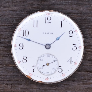 Puede incluir: Un reloj de bolsillo blanco con agujas negras y un peque&ntilde;o subdial. La esfera del reloj tiene n&uacute;meros negros y la palabra "ELGIN" impresa en ella.