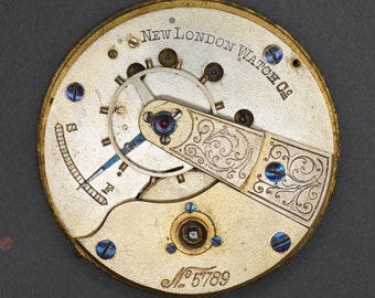 Op de afbeelding: Close-up van een goudkleurig zakhorlogemechanisme met de inscriptie "New London Watch Co." en het serienummer "#5789". Het mechanisme heeft een decoratief, gegraveerd ontwerp en blauwe accenten.