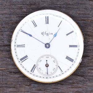 May include: A white pocket watch face with black Roman numeral hour markers and black hands. The watch face has a small seconds dial and the word "Elgin" printed in black.