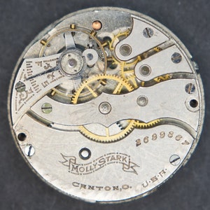 May include: Close-up of the inner workings of a silver pocket watch with gold gears. The watch face is marked "Molly Stark" and "Canton, O. U.S.A." with the serial number "2699869".