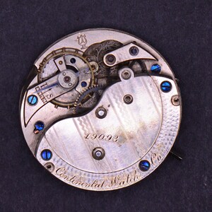 May include: Close-up of the back of a silver pocket watch with a detailed mechanical movement. The watch face has the inscription "Continental Watch Co." and the number "19093".