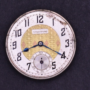 Pu&ograve; includere: Un orologio da tasca argentato con un quadrante bianco e indici orari romani neri. L'orologio ha un cerchio interno dorato con un motivo a rombi e la parola "STRATFORD" stampata in nero. L'orologio ha una lancetta blu e un quadrante bianco pi&ugrave; piccolo con segni neri per i secondi.