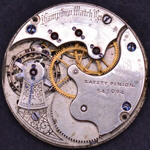 K&ouml;nnte beinhalten: Nahaufnahme eines silbernen Taschenuhrwerks mit goldenen Zahnr&auml;dern. Die Uhr ist mit "Hamden Watch Co." und "SAFETY PINION 543094" beschriftet.