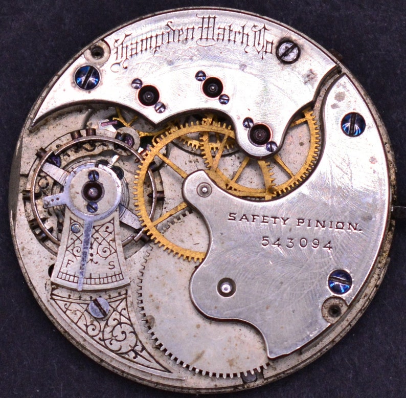 K&ouml;nnte beinhalten: Nahaufnahme eines silbernen Taschenuhrwerks mit goldenen Zahnr&auml;dern. Die Uhr ist mit "Hamden Watch Co." und "SAFETY PINION 543094" beschriftet.