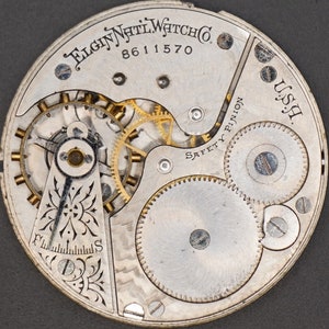 Op de afbeelding: Close-up van een zilveren zakhorloge-uurwerk met de tekst "Elgin Natl. Watch Co. 8611570 U.S.A." en "Safety Pinion". Het uurwerk heeft een decoratief bloemmotief en verschillende tandwielen.