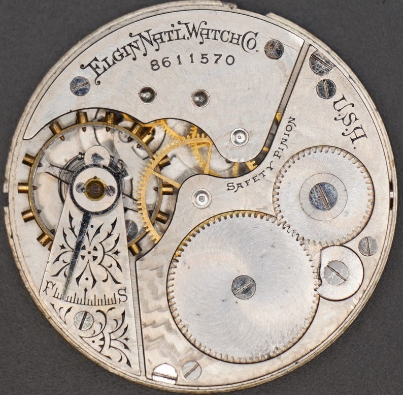 Op de afbeelding: Close-up van een zilveren zakhorloge-uurwerk met de tekst "Elgin Natl. Watch Co. 8611570 U.S.A." en "Safety Pinion". Het uurwerk heeft een decoratief bloemmotief en verschillende tandwielen.