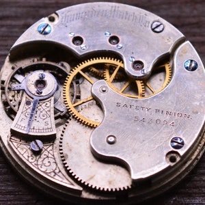 K&ouml;nnte beinhalten: Nahaufnahme eines silbernen Taschenuhrwerks mit goldenen Zahnr&auml;dern. Die Uhr hat ein Sicherheitszahnrad und die Nummer 543094 ist sichtbar. Die Uhr ist mit " Hampden Watch Co." graviert.