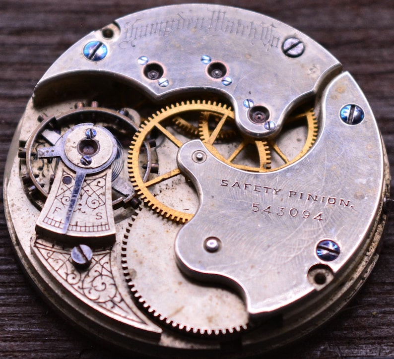 K&ouml;nnte beinhalten: Nahaufnahme eines silbernen Taschenuhrwerks mit goldenen Zahnr&auml;dern. Die Uhr hat ein Sicherheitszahnrad und die Nummer 543094 ist sichtbar. Die Uhr ist mit " Hampden Watch Co." graviert.