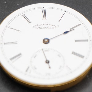 Puede incluir: Una esfera de reloj de bolsillo blanca con marcadores de hora en n&uacute;meros romanos y agujas negras. La esfera del reloj tiene el texto "American Waltham" impreso en ella.