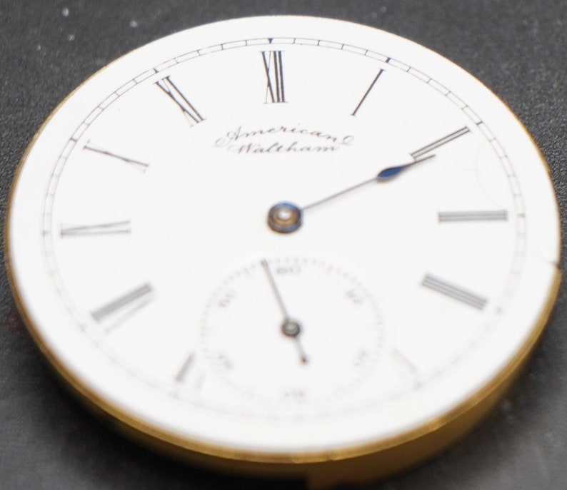 Puede incluir: Una esfera de reloj de bolsillo blanca con marcadores de hora en n&uacute;meros romanos y agujas negras. La esfera del reloj tiene el texto "American Waltham" impreso en ella.