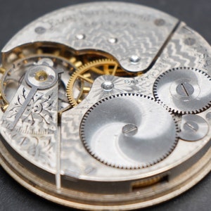 Op de afbeelding: Close-up van een zilveren zakhorloge-uurwerk met ingewikkelde tandwielen en het woord "SAFETY" gegraveerd op de wijzerplaat.