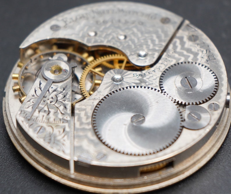 Op de afbeelding: Close-up van een zilveren zakhorloge-uurwerk met ingewikkelde tandwielen en het woord "SAFETY" gegraveerd op de wijzerplaat.