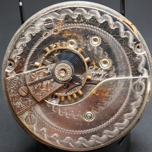 May include: Close-up of a silver pocket watch movement with intricate engravings and the text "Safety Pinion".