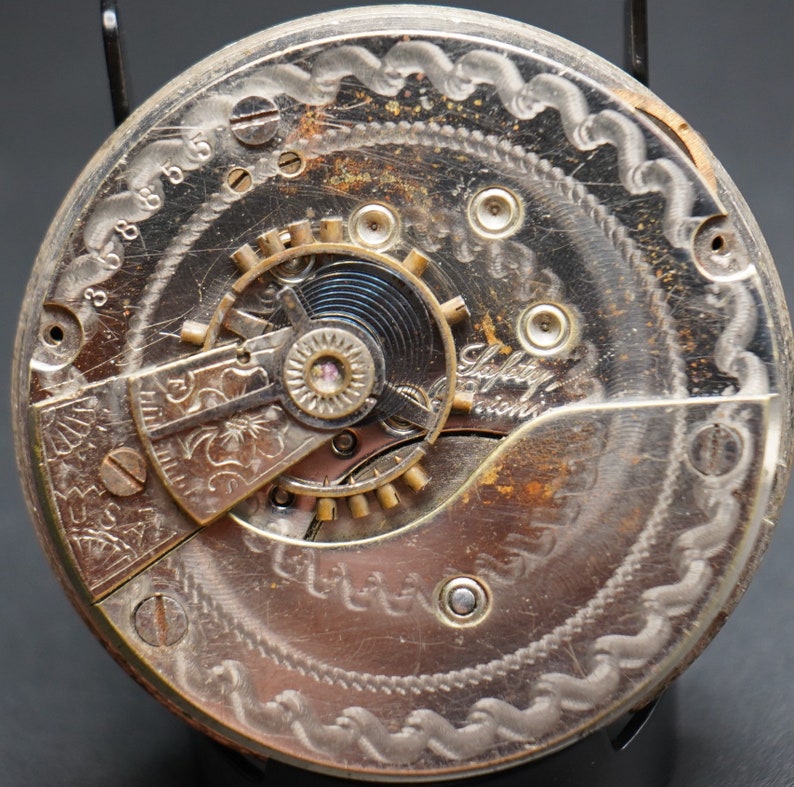 May include: Close-up of a silver pocket watch movement with intricate engravings and the text "Safety Pinion".