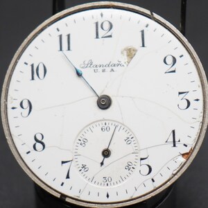 May include: A white pocket watch with black Roman numeral hour markers and a smaller subdial. The watch face is cracked and the watch is labeled "Standard U.S.A."