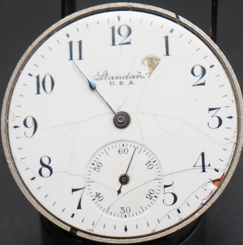 May include: A white pocket watch with black Roman numeral hour markers and a smaller subdial. The watch face is cracked and the watch is labeled "Standard U.S.A."
