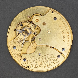 Puede incluir: Primer plano de un movimiento de reloj de bolsillo de color dorado con la inscripci&oacute;n "SAFETY BARREL 5166307" y "AMERICAN WALTHAM WATCH CO."